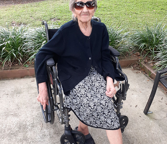 Elderly woman in wheelchair wearing sunglasses and patterned dress, outdoors with greenery in the background.