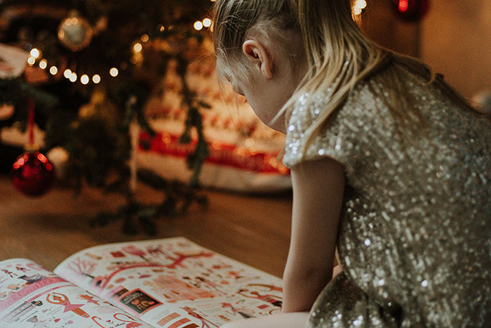 Young girl in a sparkly dress by a Christmas tree looking at a gift catalog representing worst gifts received stories.