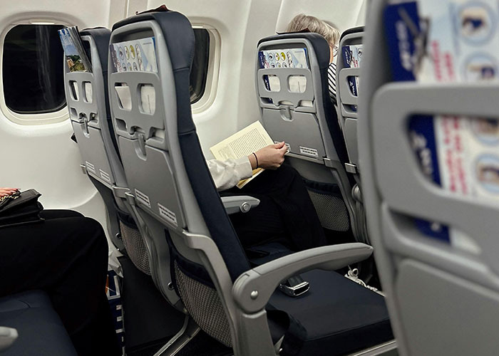 Passenger reading a book while occupying multiple airplane seats, illustrating entitled behavior on a flight in 2025.