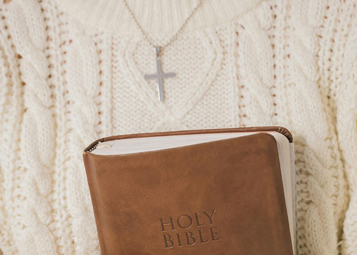 Person wearing a silver cross necklace and cream sweater holding a brown leather Holy Bible, representing entitlement concept.