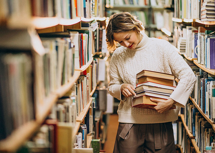 Young woman carrying a stack of books in a library aisle, unrelated to a gym membership gift concept.