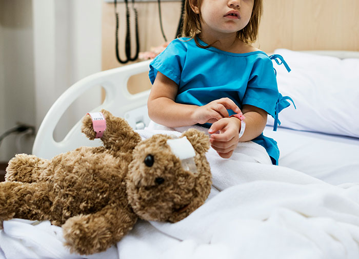 Young patient in hospital gown sitting on bed with a bandaged teddy bear, illustrating worst things patients endured.