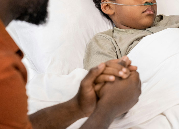 Child patient with oxygen tube lying in hospital bed while a person holds their hand showing support and care.