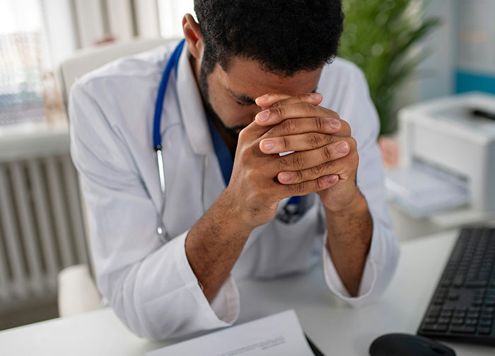 Stressed male doctor sitting at desk with hands clasped, reflecting on worst things patients had to go through in hospital.