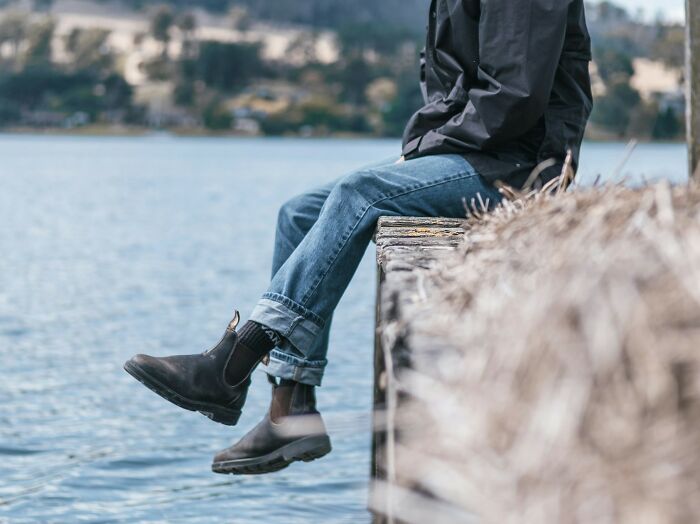 Person wearing boots and jeans sitting by the water, reflecting on mysteries that remain unsolved and puzzling.