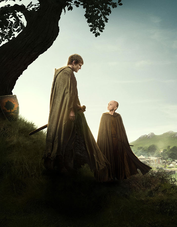 Two cloaked figures, one taller and one shorter, stand outdoors near a tree with a scenic village in the background, highlighting Peter Claffey's height.