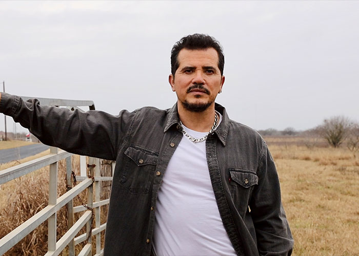 Star actor standing outdoors by a gate, wearing a dark jacket and white shirt, amid a dry grassy field under a cloudy sky.
