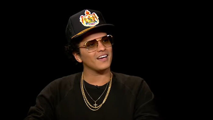 Bruno Mars wearing sunglasses and a cap, smiling during an interview about celebs who went from rags to riches.