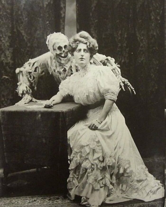 Old black and white creepy photo of a surprised woman in vintage dress with a skeleton figure behind her.