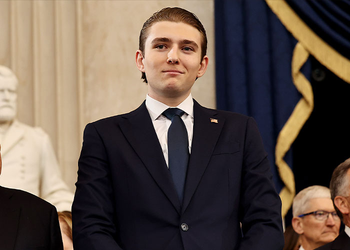 Barron Trump dressed in a suit and tie attending a formal event with a serious expression and American flag pin. Barron Trump dressed in a suit and tie attending a formal event with a serious expression and American flag pin.