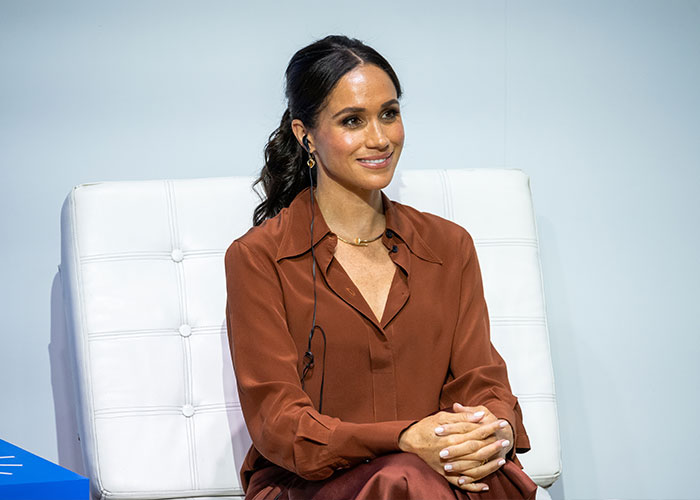 Meghan Markle seated in a brown outfit, smiling during an event, symbolizing alleged rift with mom Doria discussion.