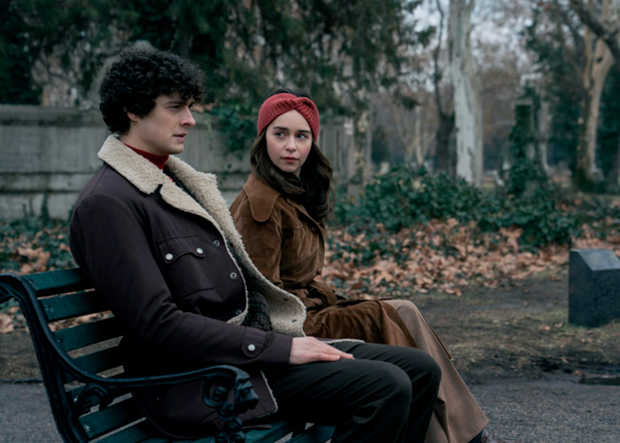 Emilia Clarke sitting on a park bench wearing a brown coat and a red headband during a film scene outdoors.