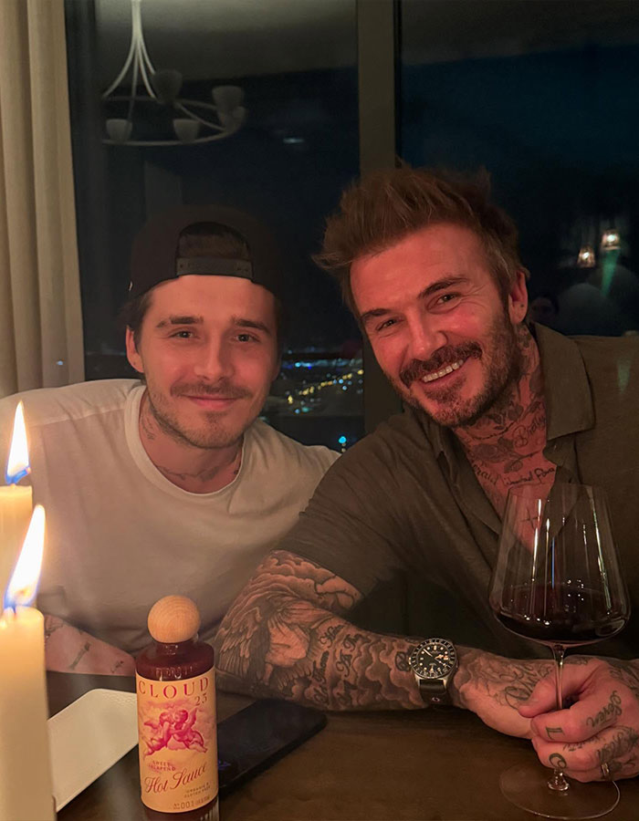 Brooklyn Beckham and David Beckham smiling at a table with candles and wine, amid family feud online discussions.