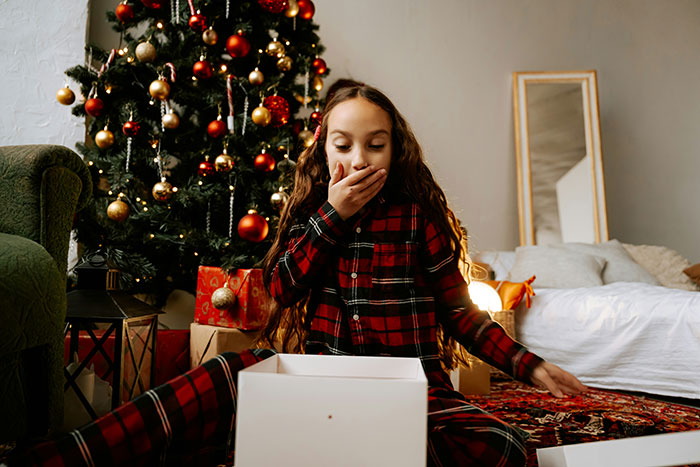 Young daughter in red pajamas surprised by Christmas presents under a decorated tree, highlighting gift imbalance with half-brother.