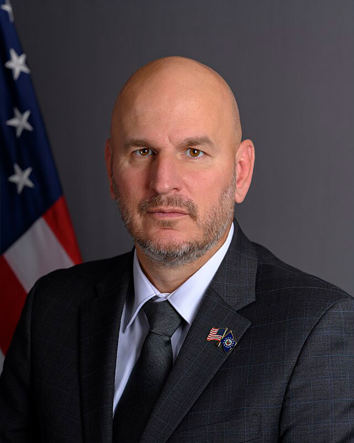 Man in dark suit with flag pins posing indoors with American flag in background, symbolizing Chile wildfires volunteer efforts. Man in dark suit with flag pins posing indoors with American flag in background, symbolizing Chile wildfires volunteer efforts.