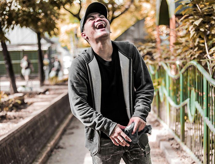 Man using theatrics to get revenge on a workplace bully, laughing outdoors with trees and fence in background