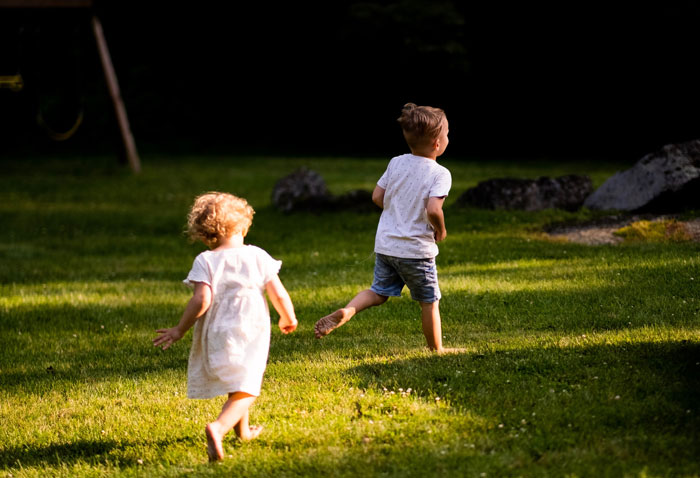 Two barefoot children running on a sunlit lawn in a backyard, playful scene, friends family weird