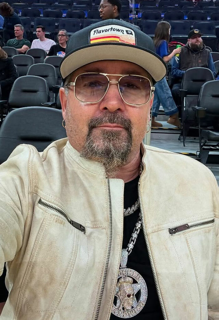 Man wearing glasses, a Flavortown cap, and a white jacket sitting among empty seats, reflecting on movie set workers and diva stars.
