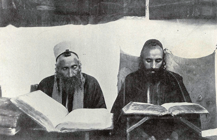 Two men with beards and traditional headwear reading large open books in an old room, representing oldest countries history.