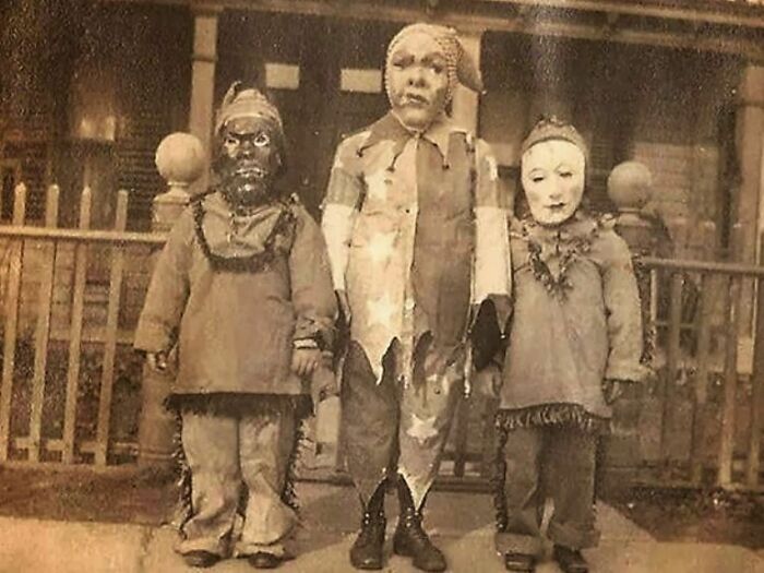 Three children in old black and white creepy costumes standing on a porch in a vintage odd and eerie photo.