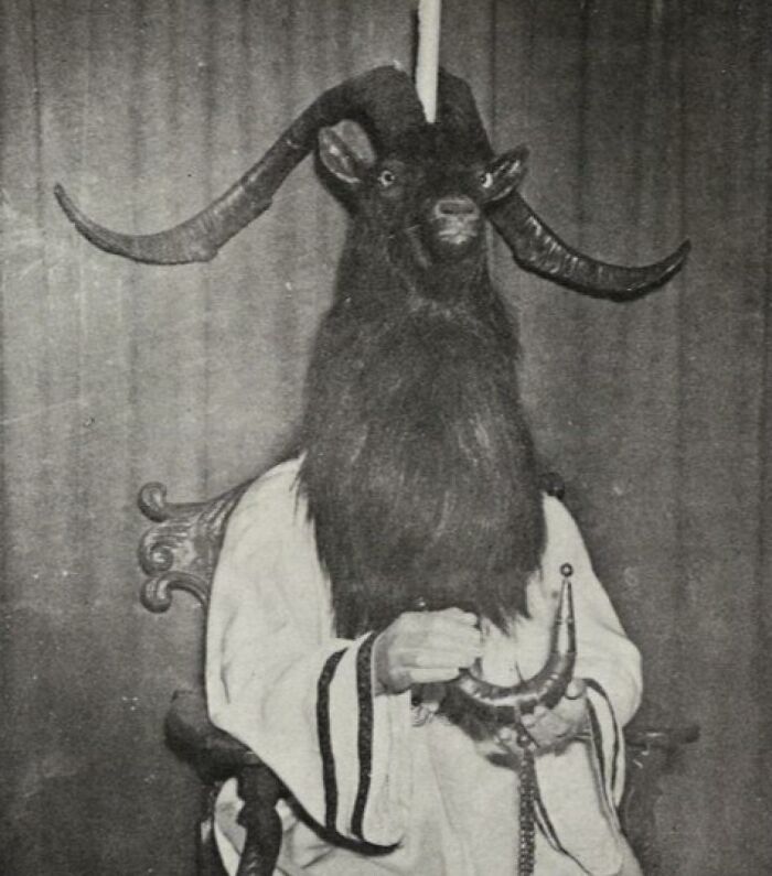 Old black and white creepy photo of a person wearing a goat head mask with large curved horns seated on a chair.