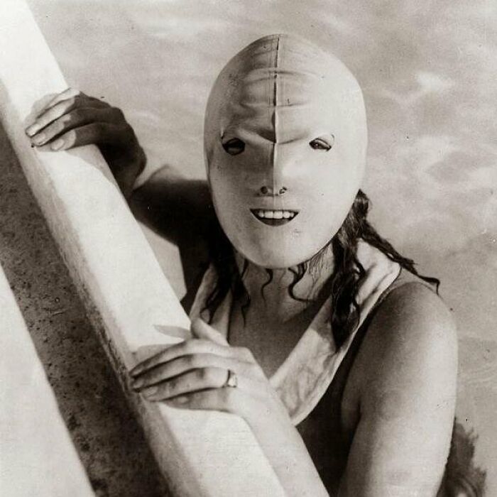 Person in old black and white photo wearing a creepy mask with eye and mouth holes while holding onto a pool edge.
