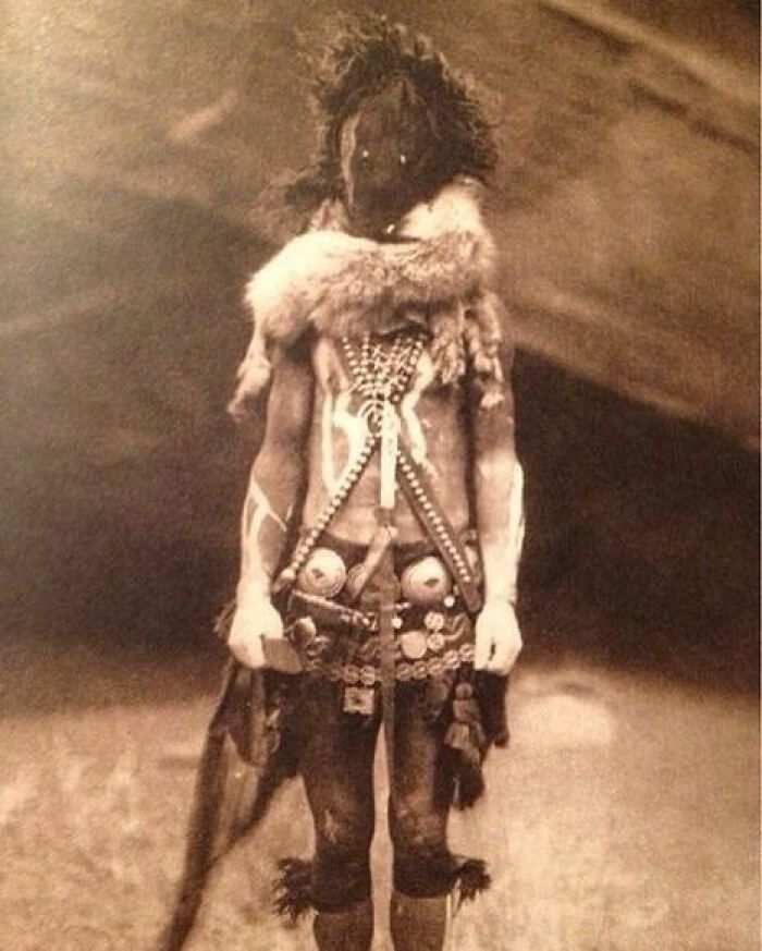 Old black and white creepy photo of a person in tribal costume with body paint and animal fur accessories.