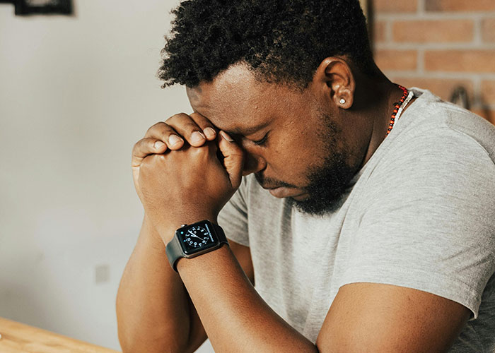 Man wearing gray shirt and smartwatch, sitting with hands clasped and eyes closed, reflecting on fatherhood and biological parenthood.