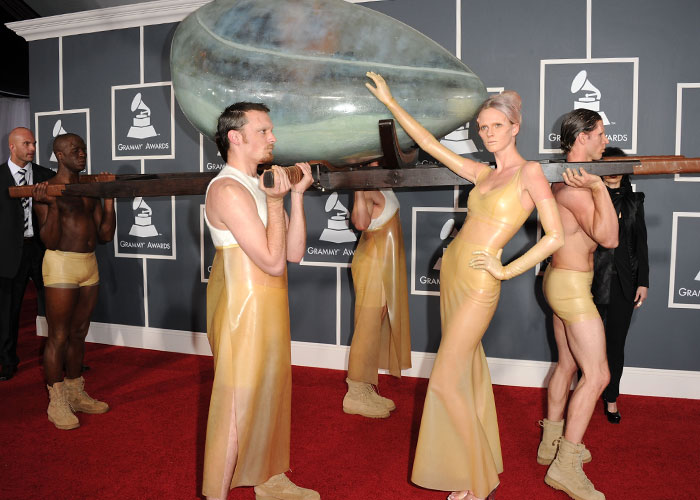 Models in avant-garde latex outfits carrying an oversized spoon prop at a wild Grammys fashion event.
