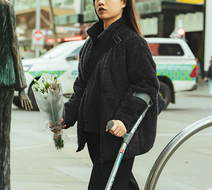 Young woman with crutch holding flowers on city street, highlighting coworker filming to prove disability claim.