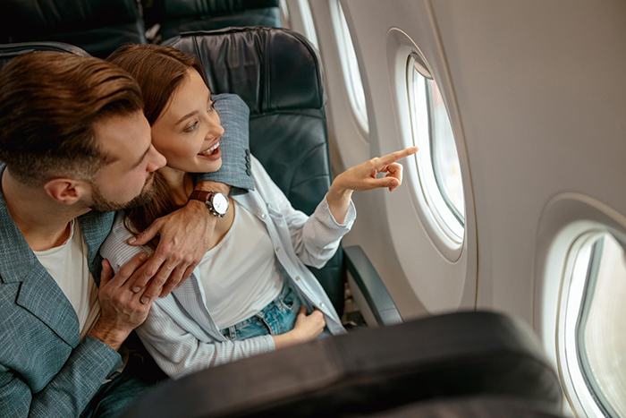 Couple seated on a plane, woman points out the window while man looks on, illustrating guy&rsquo;s heroic actions theme.