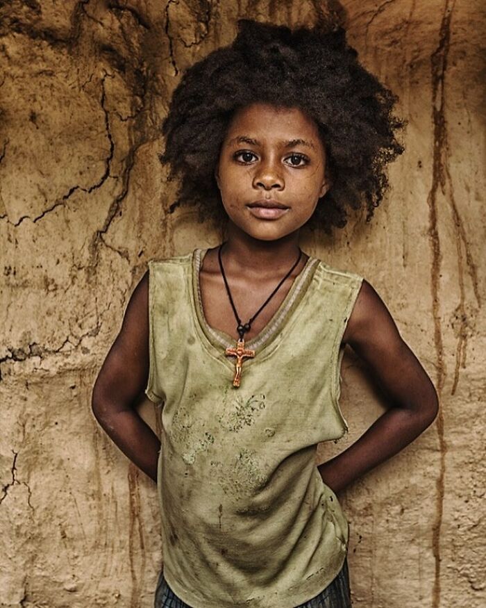 Candid street photo of a young child with natural hair standing against a textured wall, capturing raw humanity and emotion.