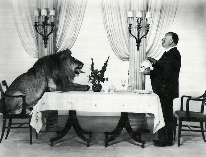 Old black and white photo of a man holding a skull while a lion sits at a dining table in a vintage room.