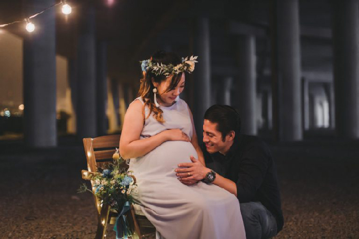 Nighttime creative maternity photoshoot with expectant mother in floral crown seated, partner kneeling and touching her belly