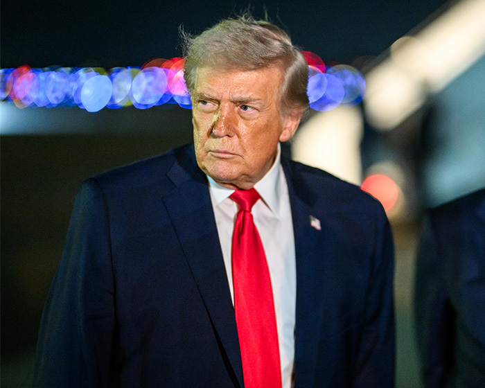 Donald Trump in a dark suit and red tie, standing outdoors at night with blurred colorful lights in the background.