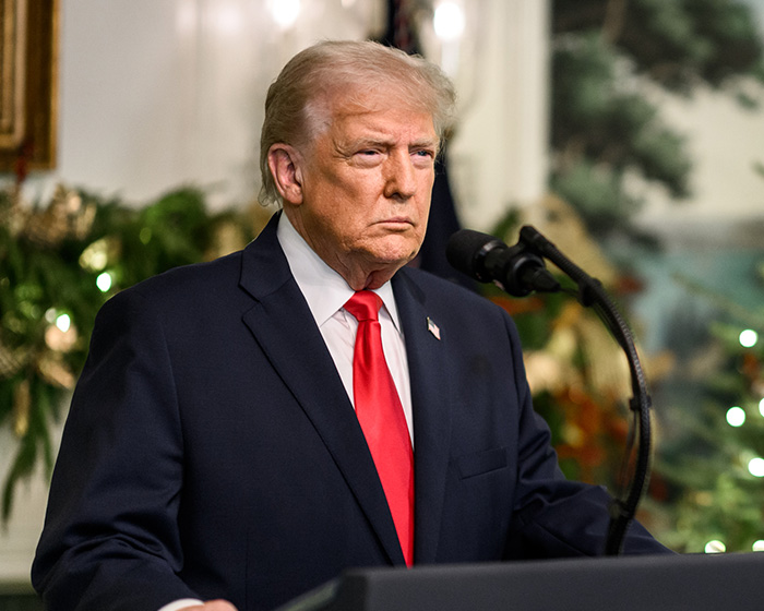 Former president Trump speaking at a podium during a formal event with holiday decorations in the background. Former president Trump speaking at a podium during a formal event with holiday decorations in the background.