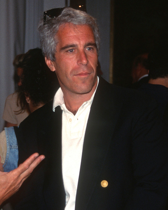 Jeffrey Epstein wearing a black blazer and white shirt at an indoor event with people in the background.