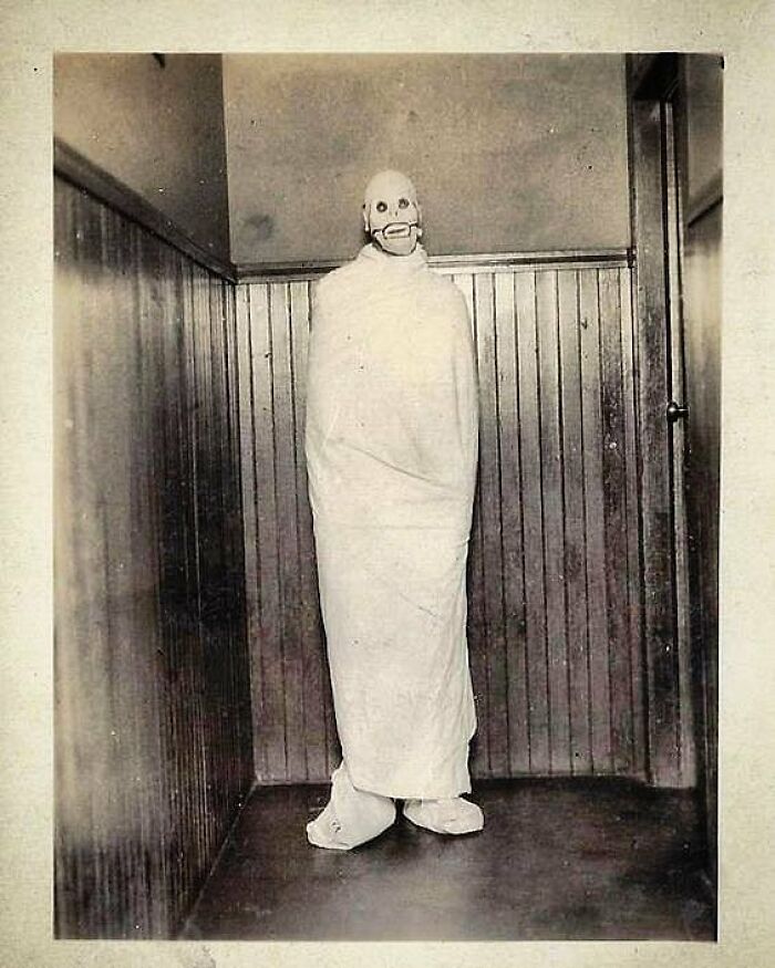 Old black and white photo of a person in a creepy mask and wrapped in cloth standing in a corner of a room.