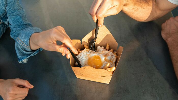 Two people sharing a meal with a fried egg on top, using forks to eat from a takeout container, debating right choices.