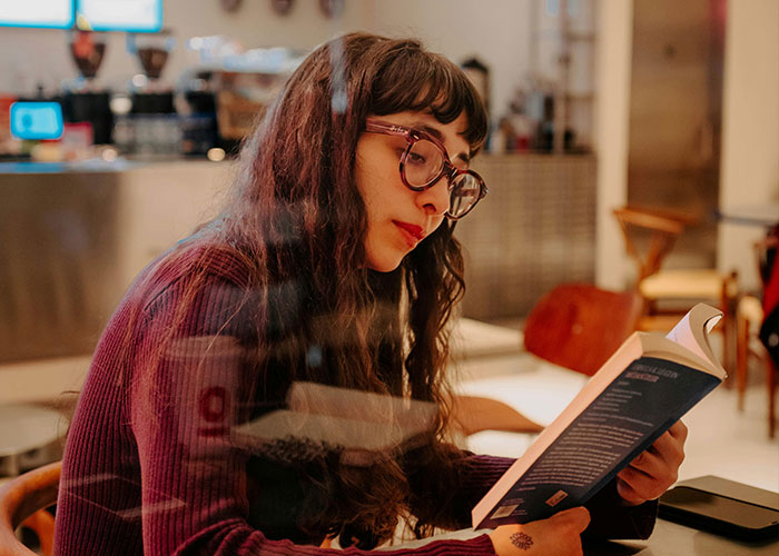 Young woman with glasses reading a book in a cafe, reflecting on experiences related to being neurodivergent.