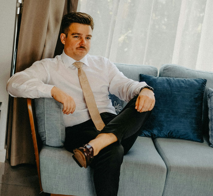 Man sitting on a couch in a white shirt and beige tie, looking thoughtful and embodying borderline smart confidence.
