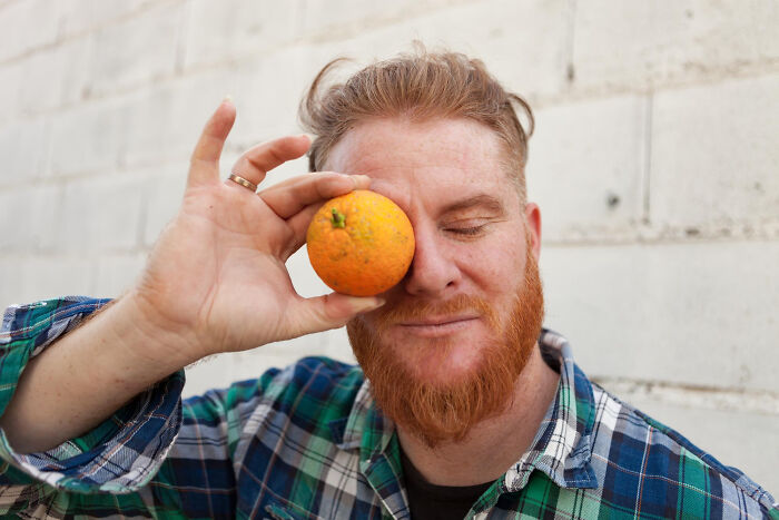 Redheaded man in plaid shirt holding an orange to his eye, capturing moments that hit people harder than expected.