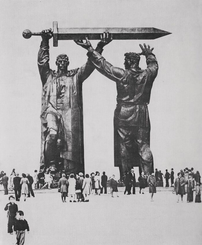 Monumental statue of two men holding a large sword with crowds gathered, reflecting the history of humanity.