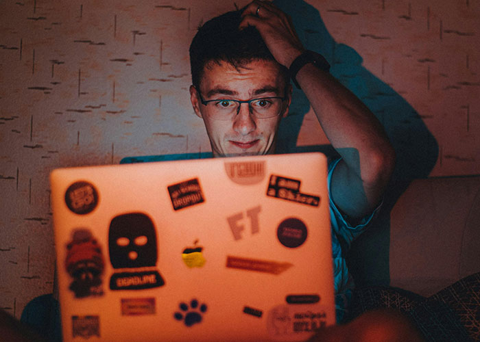 A neurodivergent person with glasses looks puzzled while using a laptop covered in various stickers in dim lighting.