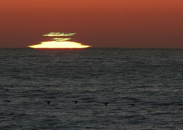 Sunset over the ocean with a wacky atmospheric illusion and birds flying low above the water surface.