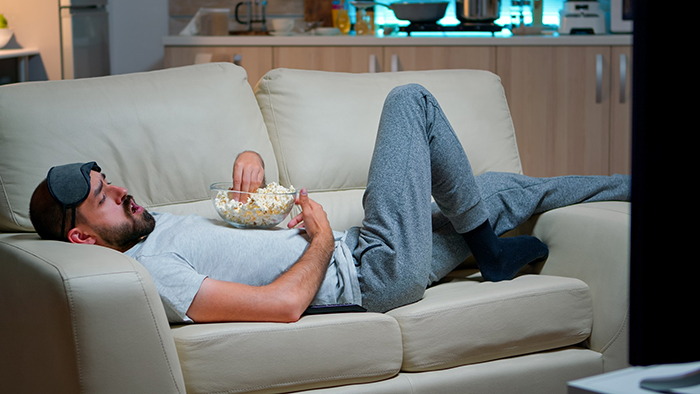 Man relaxing on couch with eye mask and popcorn, showing husband chill lifestyle in living room setting.