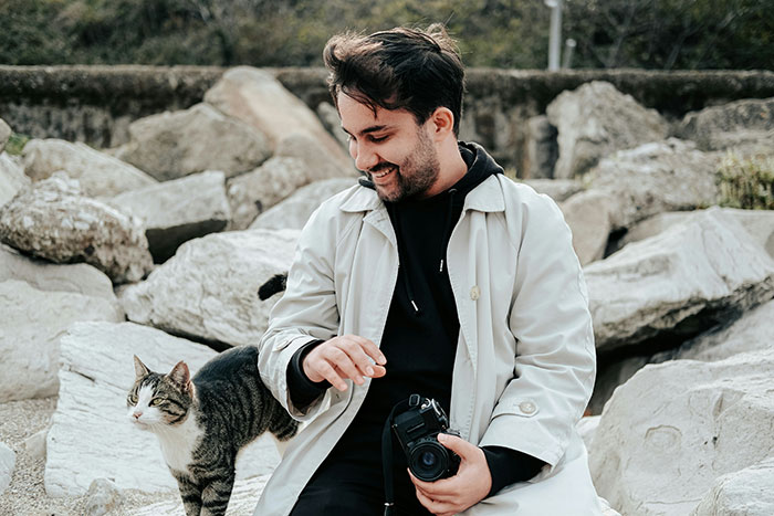 Man in light coat holding camera, smiling at a cat beside him among rocks, showing a cat disappearing and neighbor adopting theme.