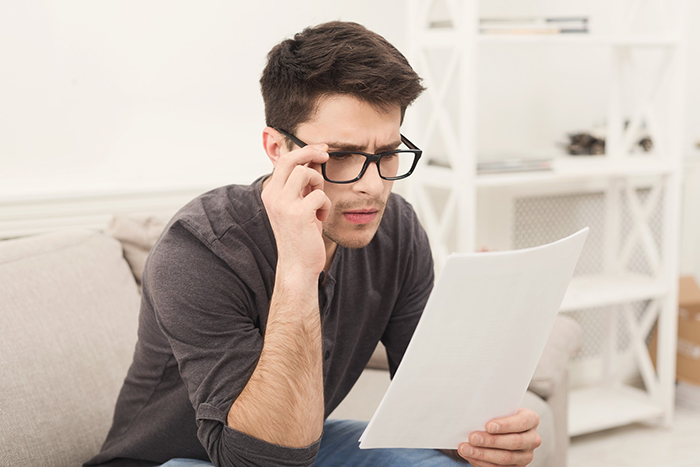 Man with glasses sitting on sofa looking surprised while reading paper about secret family discovery from ancestry test.