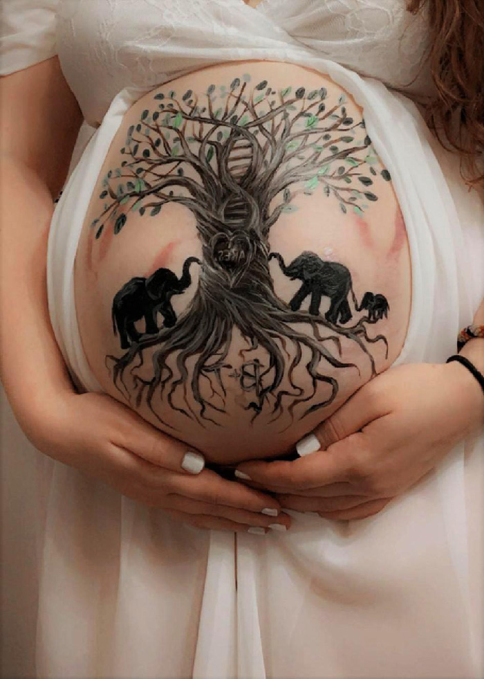 Maternity photoshoot showing painted baby bump with family tree and elephant family, hands cradling white gown.