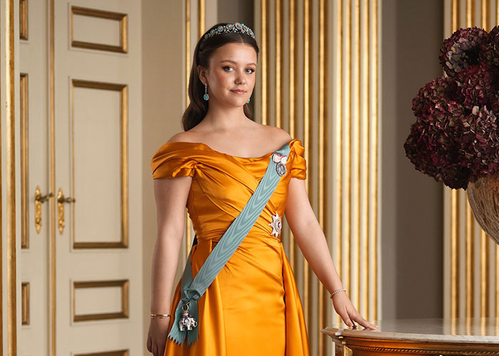 Princess Isabella wearing a golden gown and tiara, posing in an elegant room with classic decor and floral arrangement.